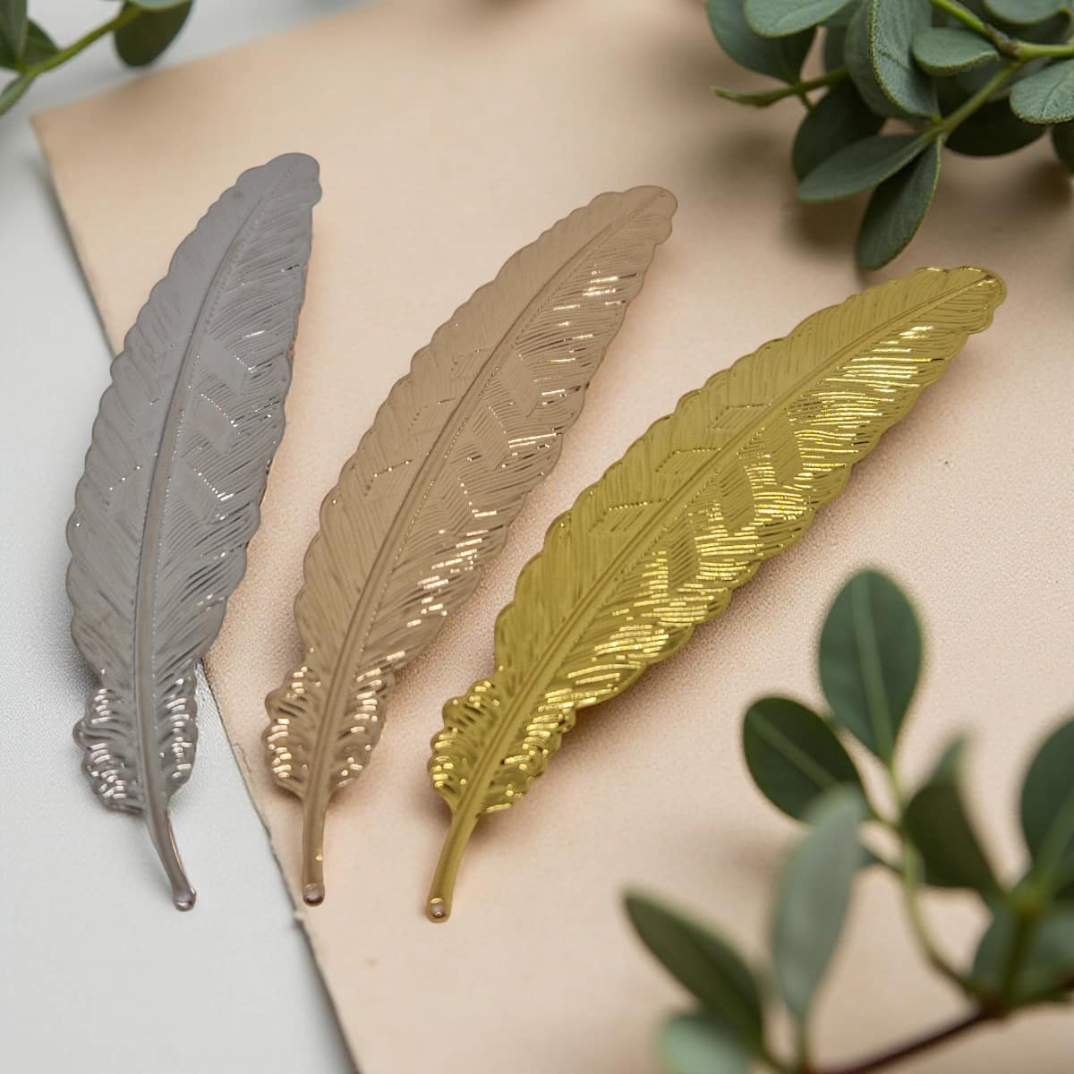 Three metallic leaf-shaped bookmark in silver, gold, and bronze on a beige surface with green leaves in the background.