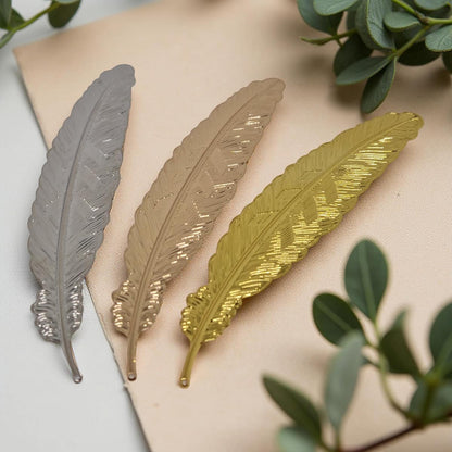 Three metallic leaf-shaped bookmark in silver, gold, and bronze on a beige surface with green leaves in the background.
