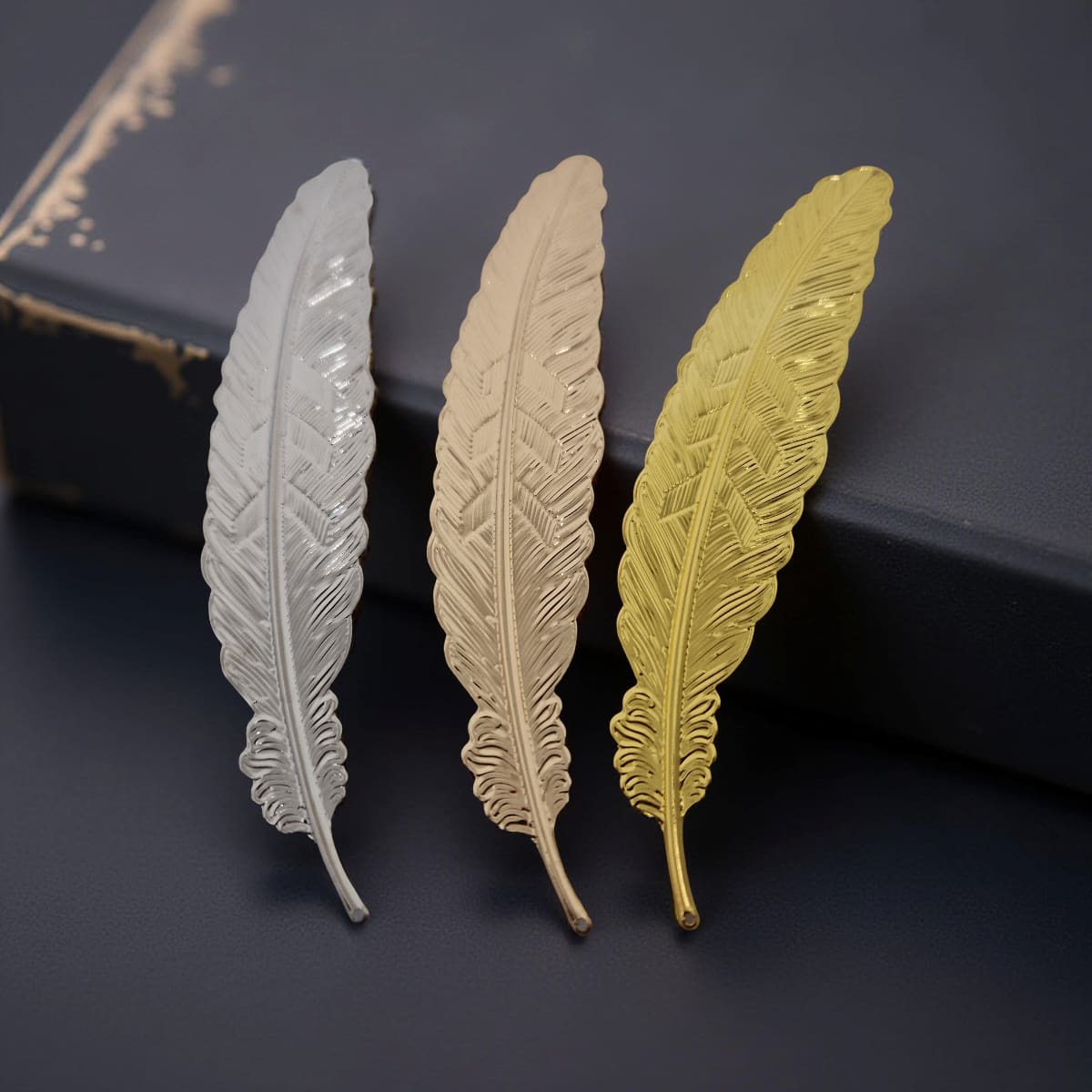 Three metallic feather-shaped bookmarks in silver, gold, and bronze on a dark surface.