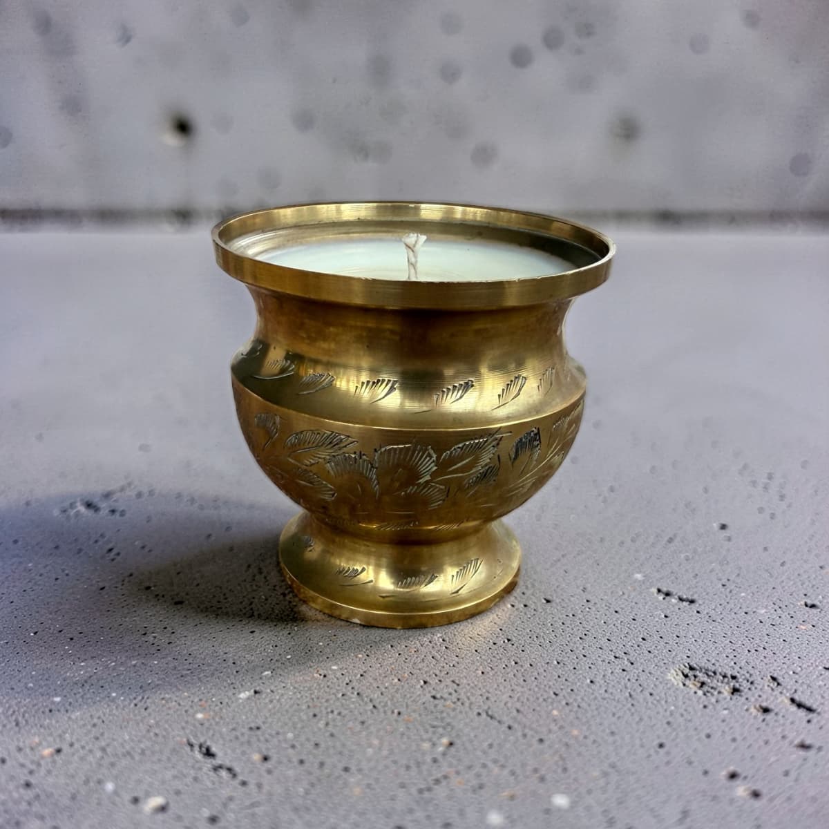 A brass candle holder with an engraved floral pattern, holding a white wax candle, set on a textured gray surface.