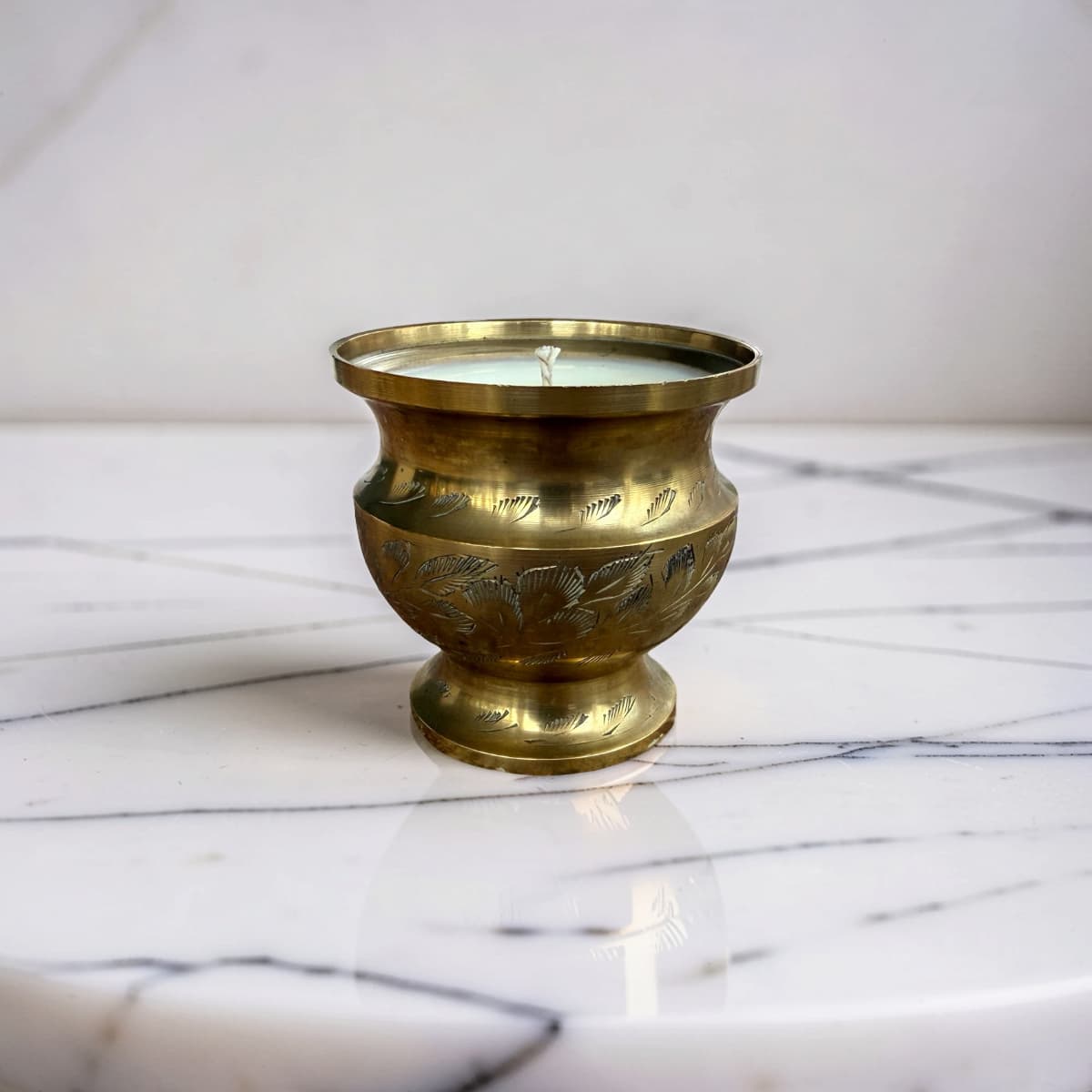 A vintage brass candle holder with engraved floral patterns, containing a white wax candle, set on a white marble surface.