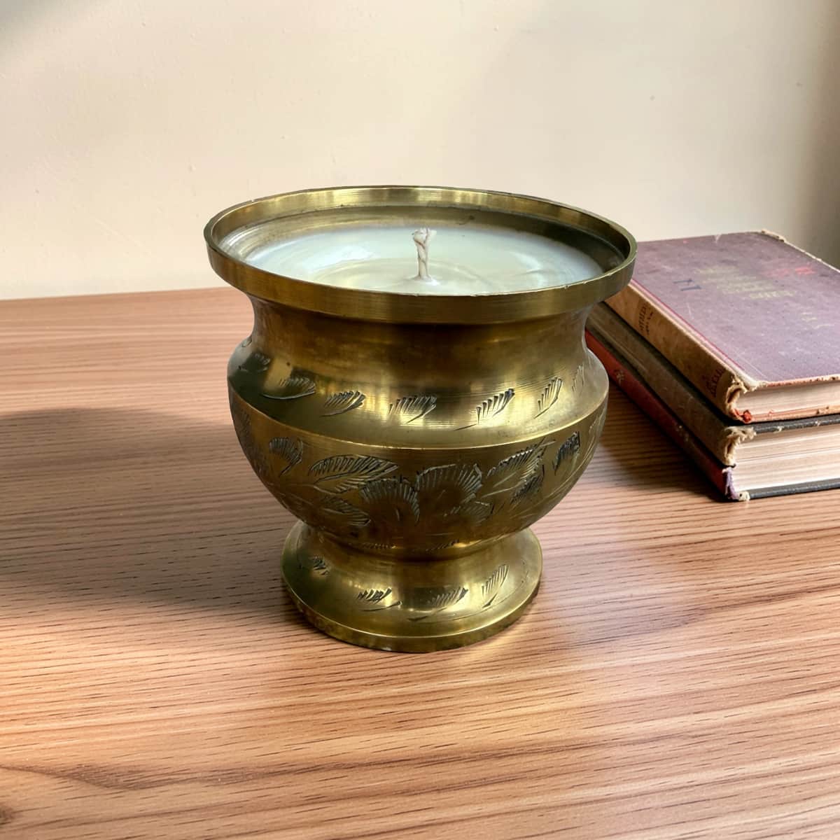 A vintage brass candle holder with a footed base and engraved floral patterns, containing a white wax candle, set on a wooden table next to a stack of old books.