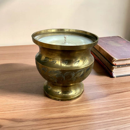 A vintage brass candle holder with a footed base and engraved floral patterns, containing a white wax candle, set on a wooden table next to a stack of old books.