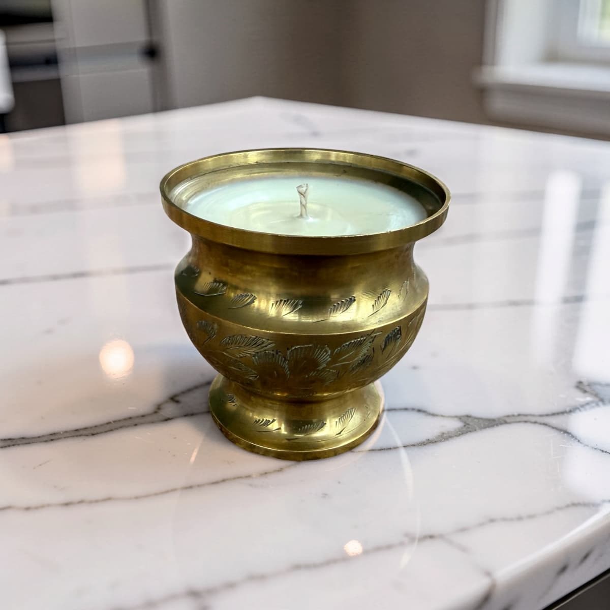 A brass candle holder with engraved leaf patterns, filled with white wax, sitting on a white marble countertop.