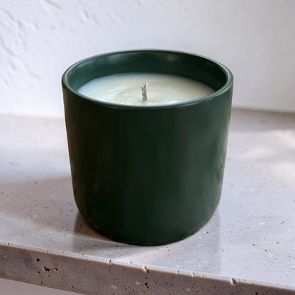 A dark green ceramic candle jar with white wax and a single wick, sitting on a speckled light gray countertop against a white textured wall.