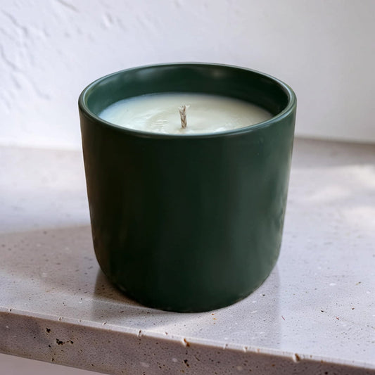 A dark green ceramic candle jar with white wax and a single wick, sitting on a speckled light gray countertop against a white textured wall.