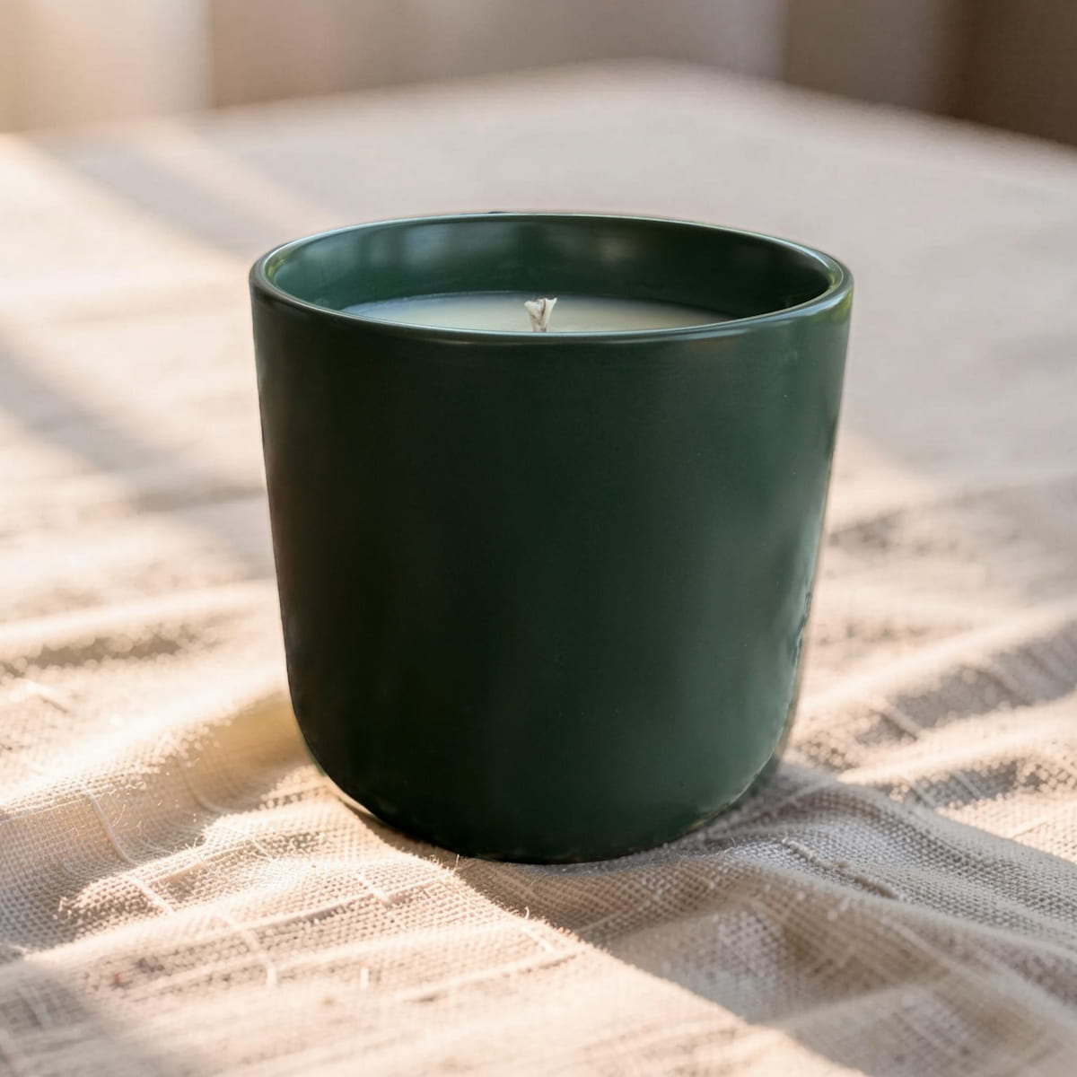 A dark green ceramic candle jar with a single wick, filled with white wax, resting on a textured beige linen cloth with soft natural light shadows.