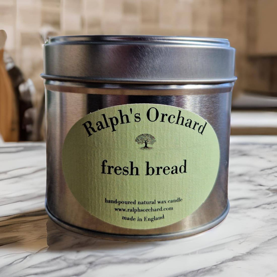 Candle in a metal tin labeled 'Ralph's Orchard fresh bread' on a marble surface.