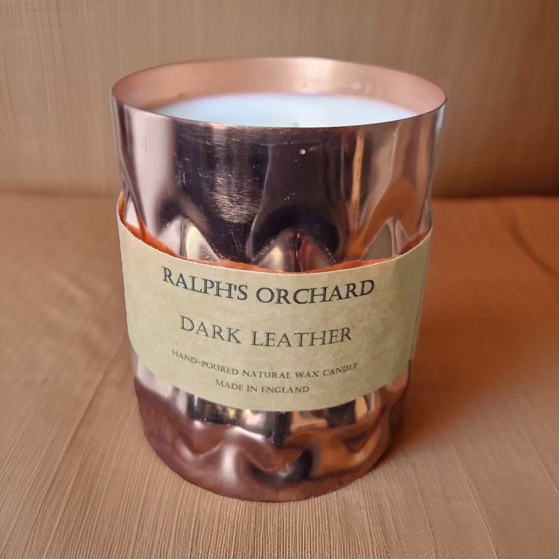 Candle in a copper holder with a label reading 'Ralph's Orchard Dark Leather' on a wooden surface.