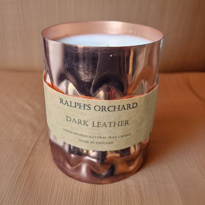 Candle in a copper holder with a label reading 'Ralph's Orchard Dark Leather' on a wooden surface.