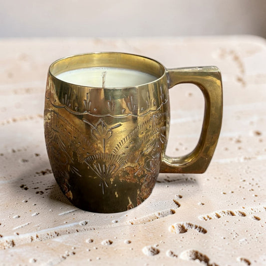 A brass mug candle with a white wax fill and a single wick, featuring hand-engraved floral and leaf patterns on the front, set on a textured beige stone surface.
