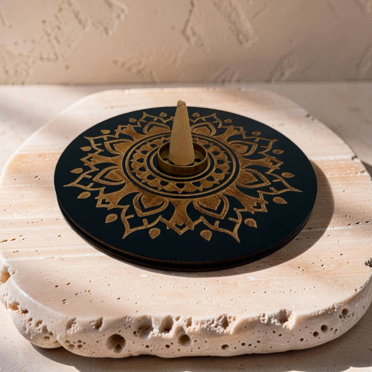 A round black and gold mandala-patterned incense cone holder or incense stand with a central cone holder, displayed on a textured beige stone slab against a beige wall.