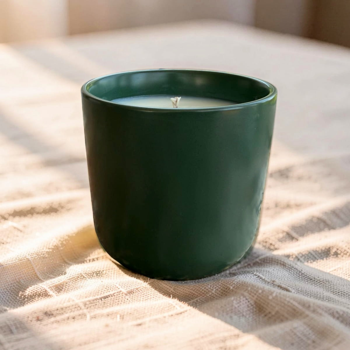 A dark green ceramic candle jar with a single wick, filled with white wax, set on a textured beige linen cloth with soft natural light.