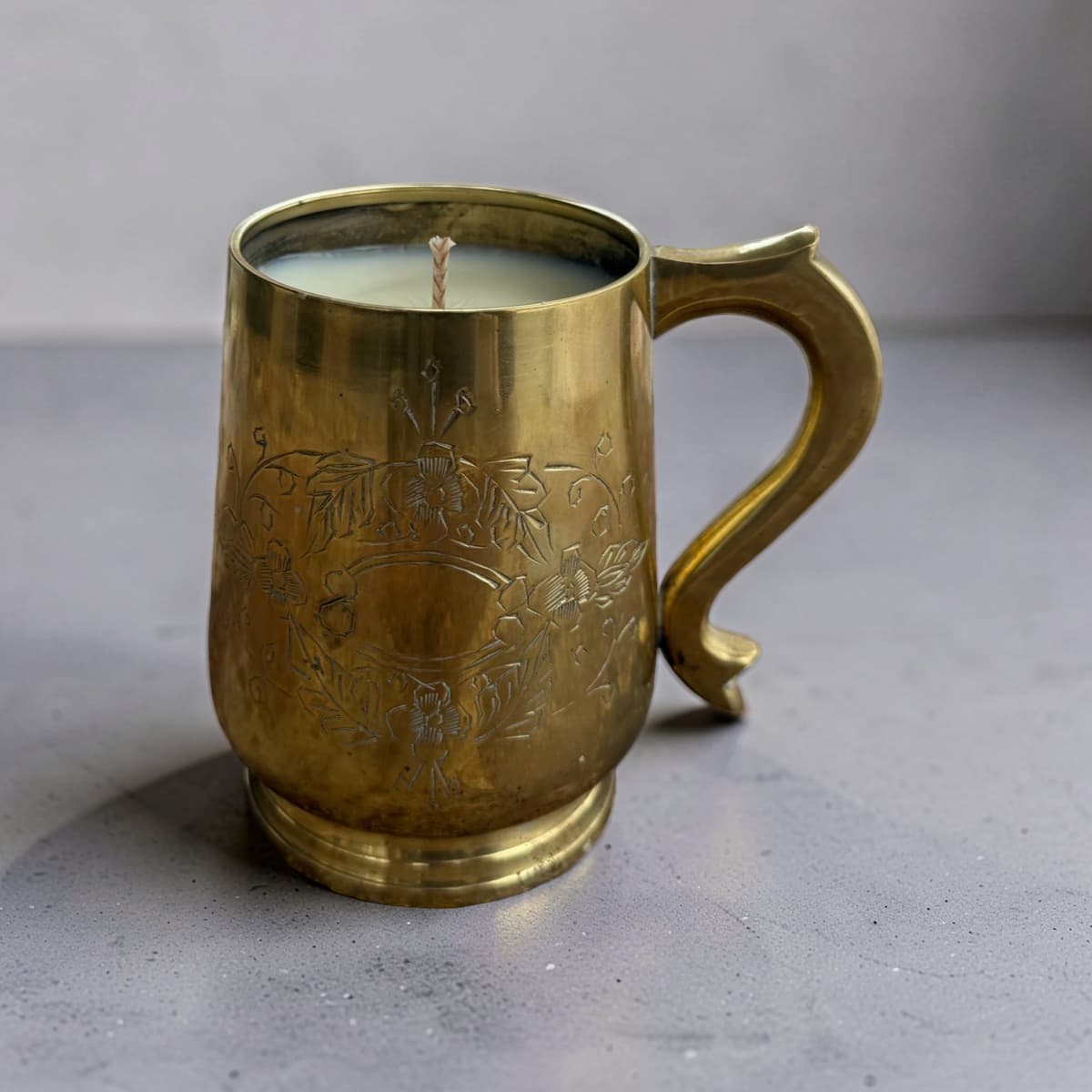 A vintage-style brass mug candle with floral engravings, filled with white wax and featuring a single wick, set against a neutral gray background.