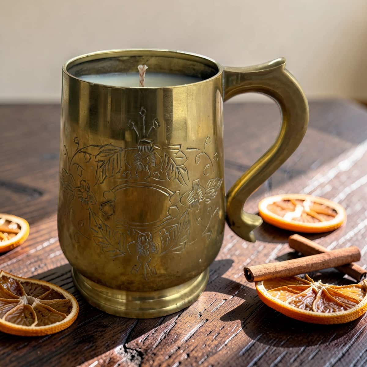 A brass mug candle with floral engravings sits on a rustic wooden table surrounded by dried orange slices, cinnamon sticks, and a few more orange slices.