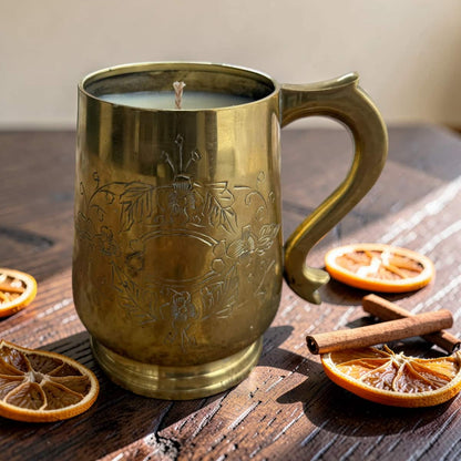 A brass mug candle with floral engravings sits on a rustic wooden table surrounded by dried orange slices, cinnamon sticks, and a few more orange slices.