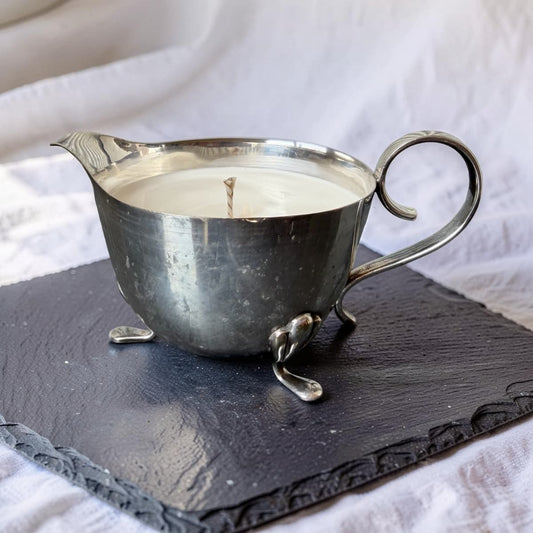 Candle in a silver pitcher on a dark slate surface with a white fabric background