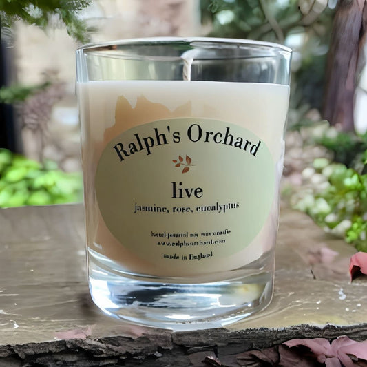 Candle in a glass jar with 'Ralph's Orchard' label on a wooden surface with blurred greenery in the background