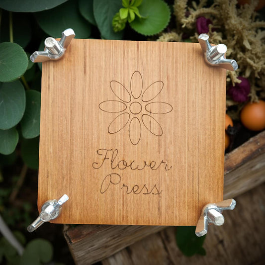 Wooden flower press with engraved design and text, surrounded by greenery.