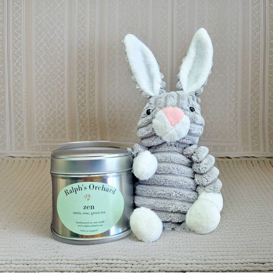 Plush rabbit toy next to a candle labeled 'Ralph's Orchard Zen' on a textured surface.