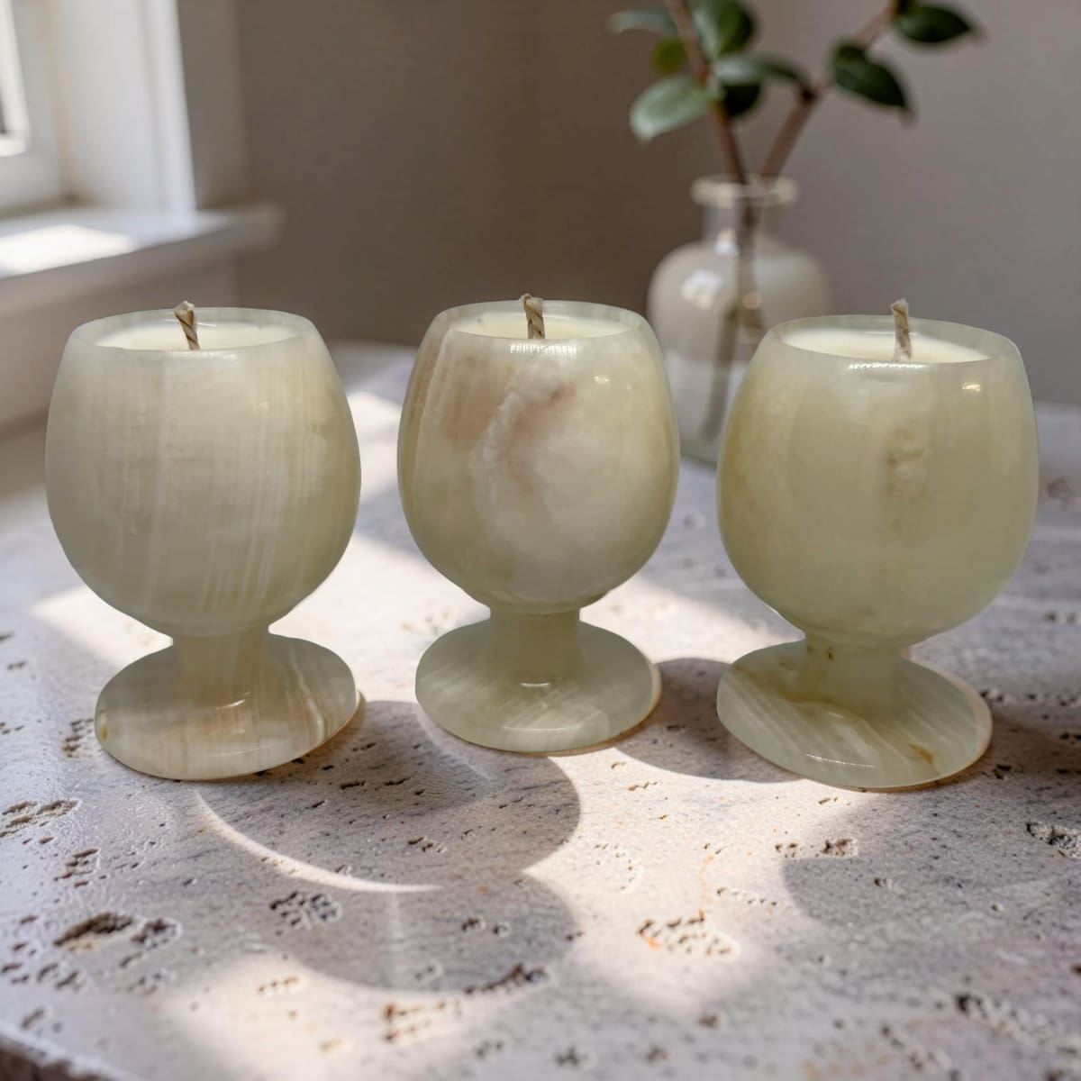 Three marble-textured candles in glass holders on a textured surface with a plant in the background.