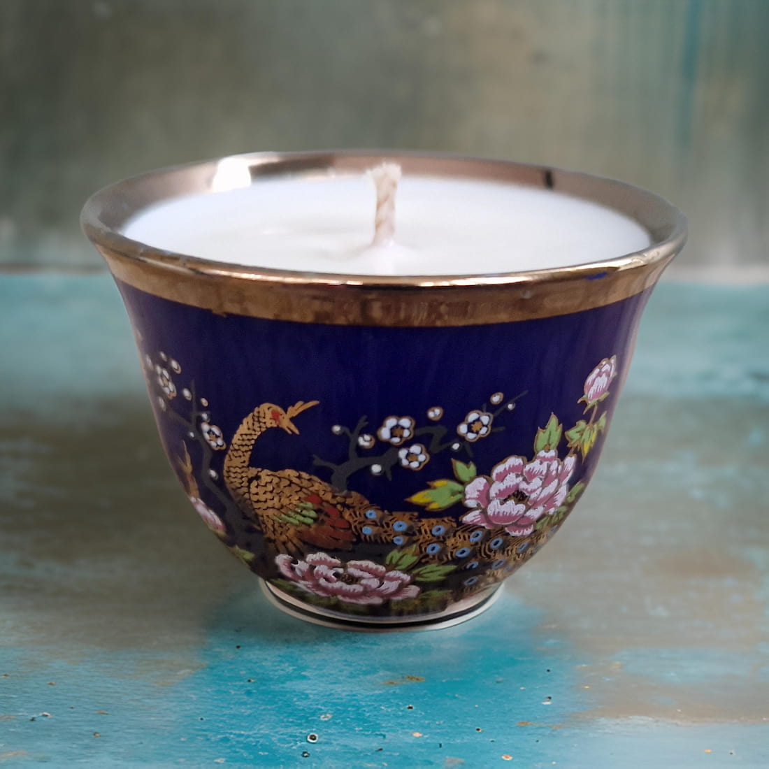 Candle in a decorative blue holder with peacock and floral design on a textured surface.