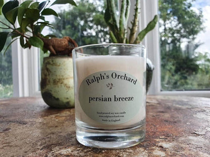 Candle labeled 'Ralph's Orchard Persian Breeze' on a wooden surface with plants in the background