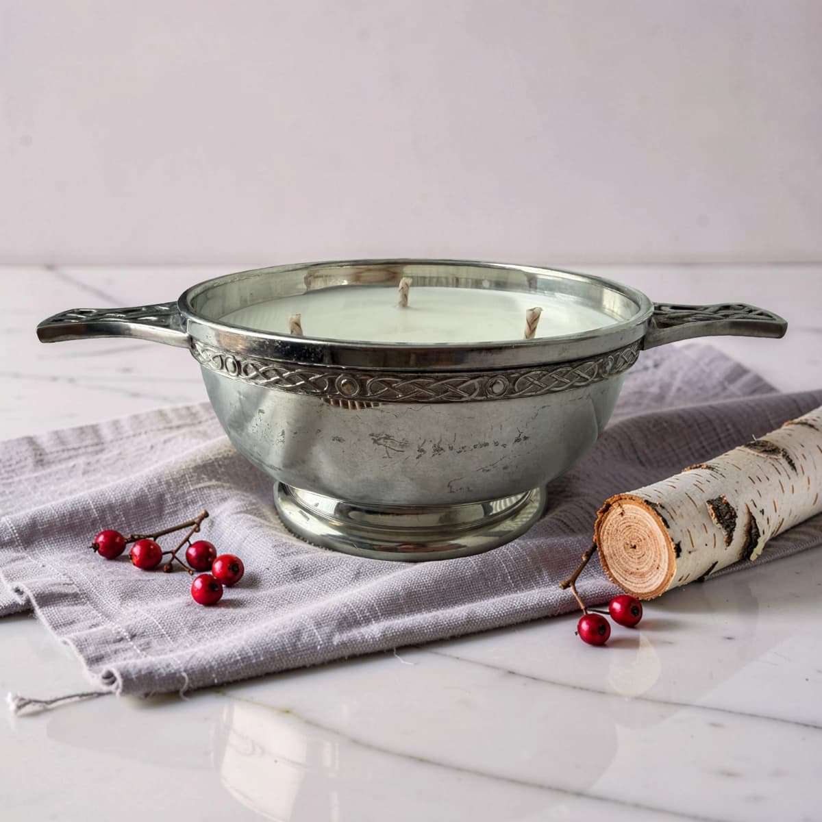 Decorative candle in a silver bowl with berries and a birch log on a white surface