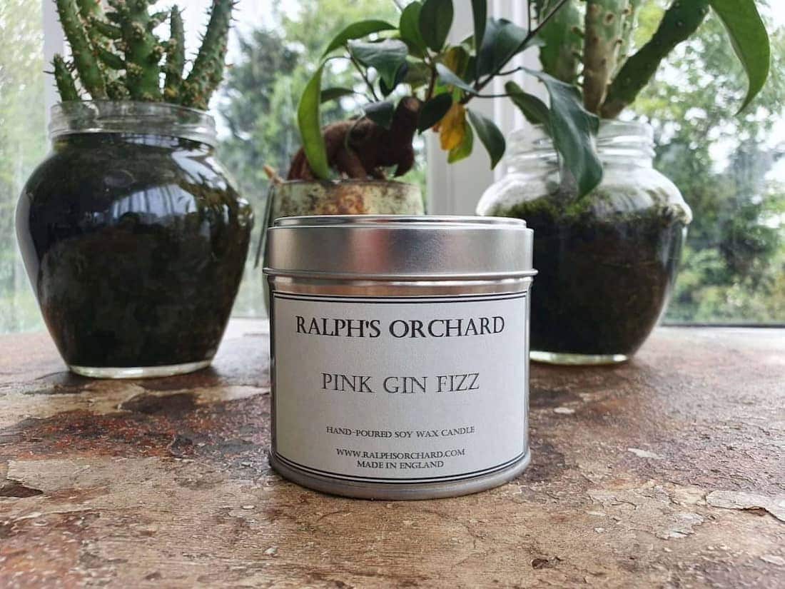 Candle labeled 'Ralph's Orchard Pink Gin Fizz' on a wooden surface with plants in the background.