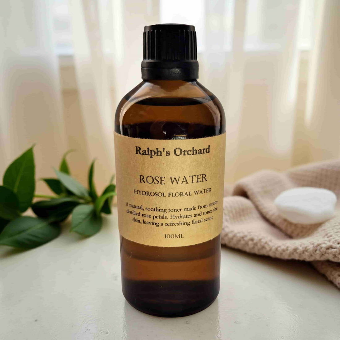 Bottle of Ralph's Orchard Rose Water on a neutral background with greenery and a towel.