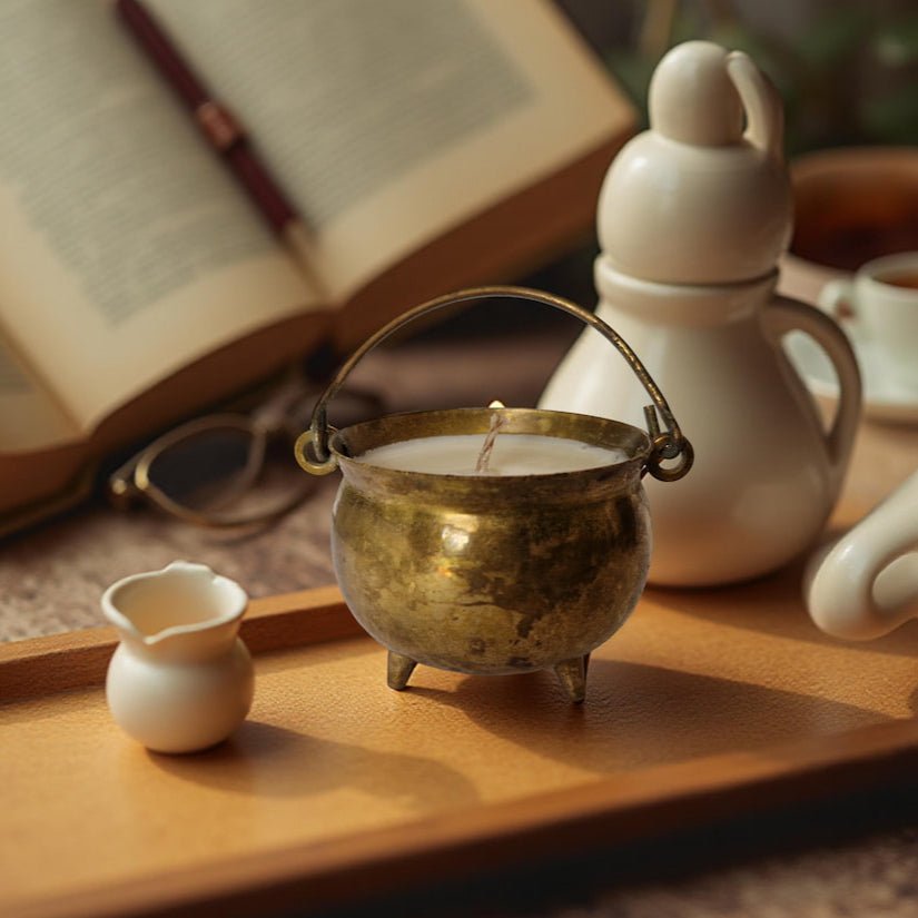 Candle in a brass holder on a wooden tray with books and ceramic items in the background