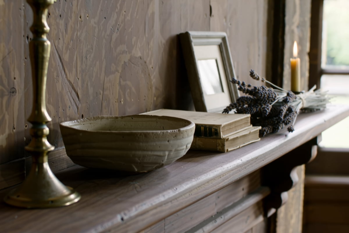 A rustic ceramic bowl with a textured, earthy finish sits on a wooden mantelpiece alongside stacked books, a small picture frame, dried lavender, and a lit candle.