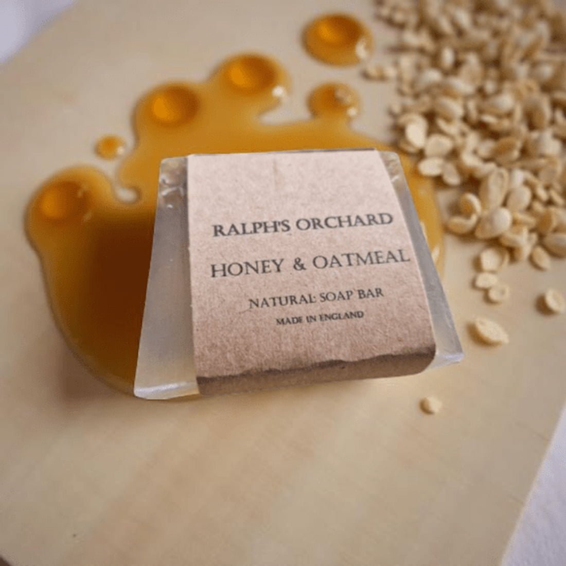 Soap bar labeled 'Ralph's Orchard Honey & Oatmeal' with honey and oats on a wooden surface.