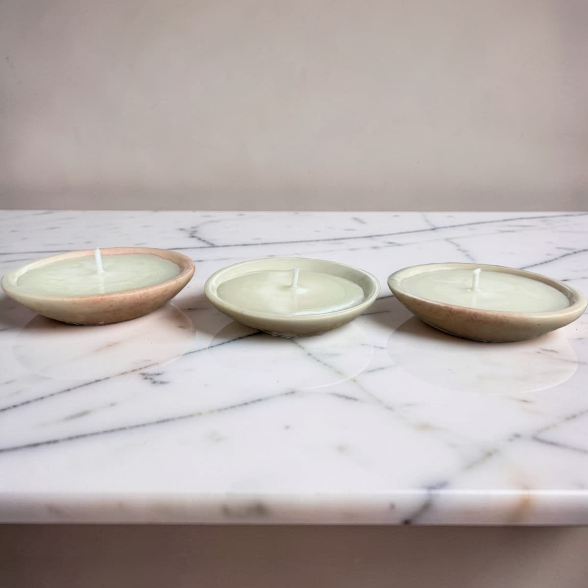Three small round candles in shallow ceramic holders, arranged side by side on a white marble surface against a plain beige wall.