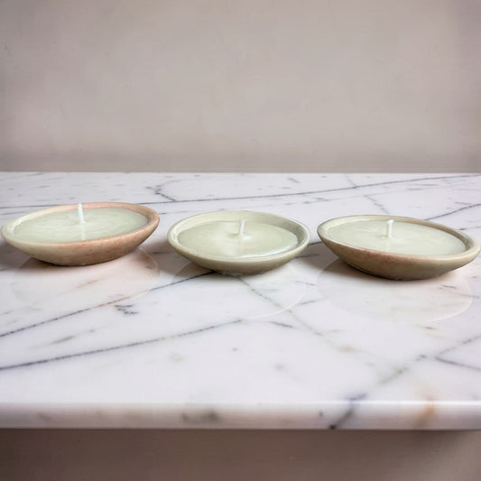 Three small round candles in shallow ceramic holders, arranged side by side on a white marble surface against a plain beige wall.