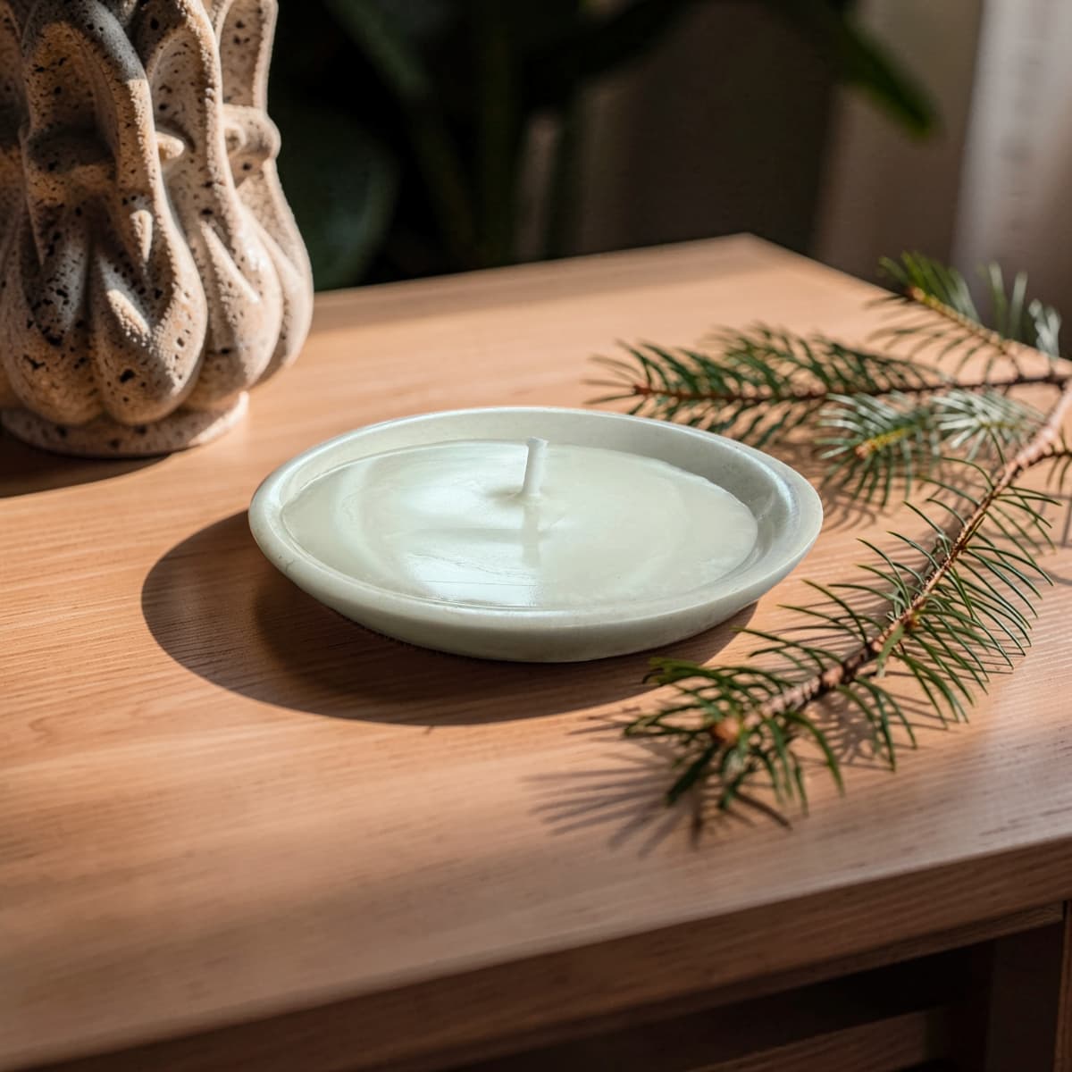 A round ceramic candle holder with a single white candle inside, placed on a light wood table next to a pine branch, with a textured ceramic vase in the background.