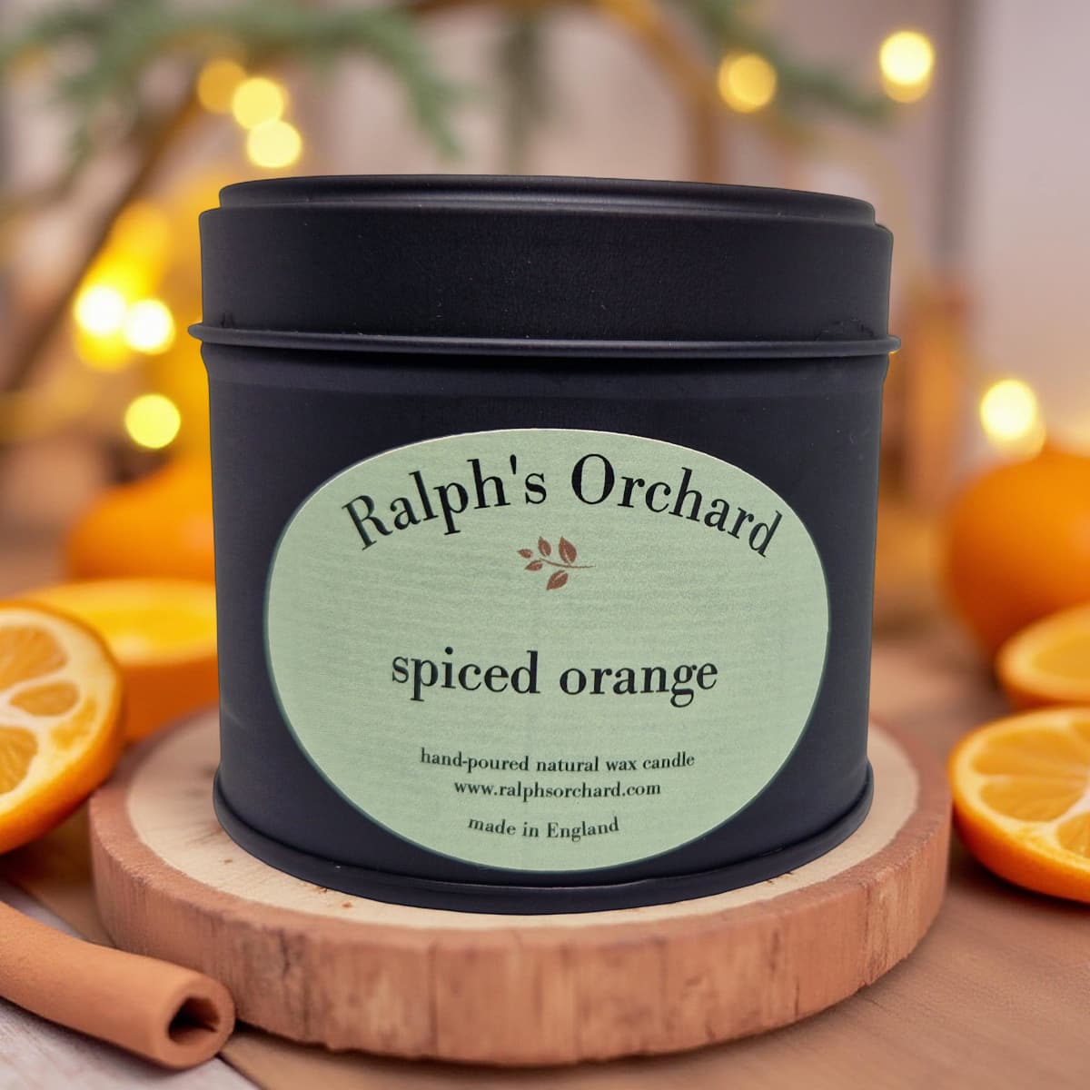 Candle in a black tin labeled 'Ralph's Orchard spiced orange' on a wooden coaster with oranges and cinnamon sticks.