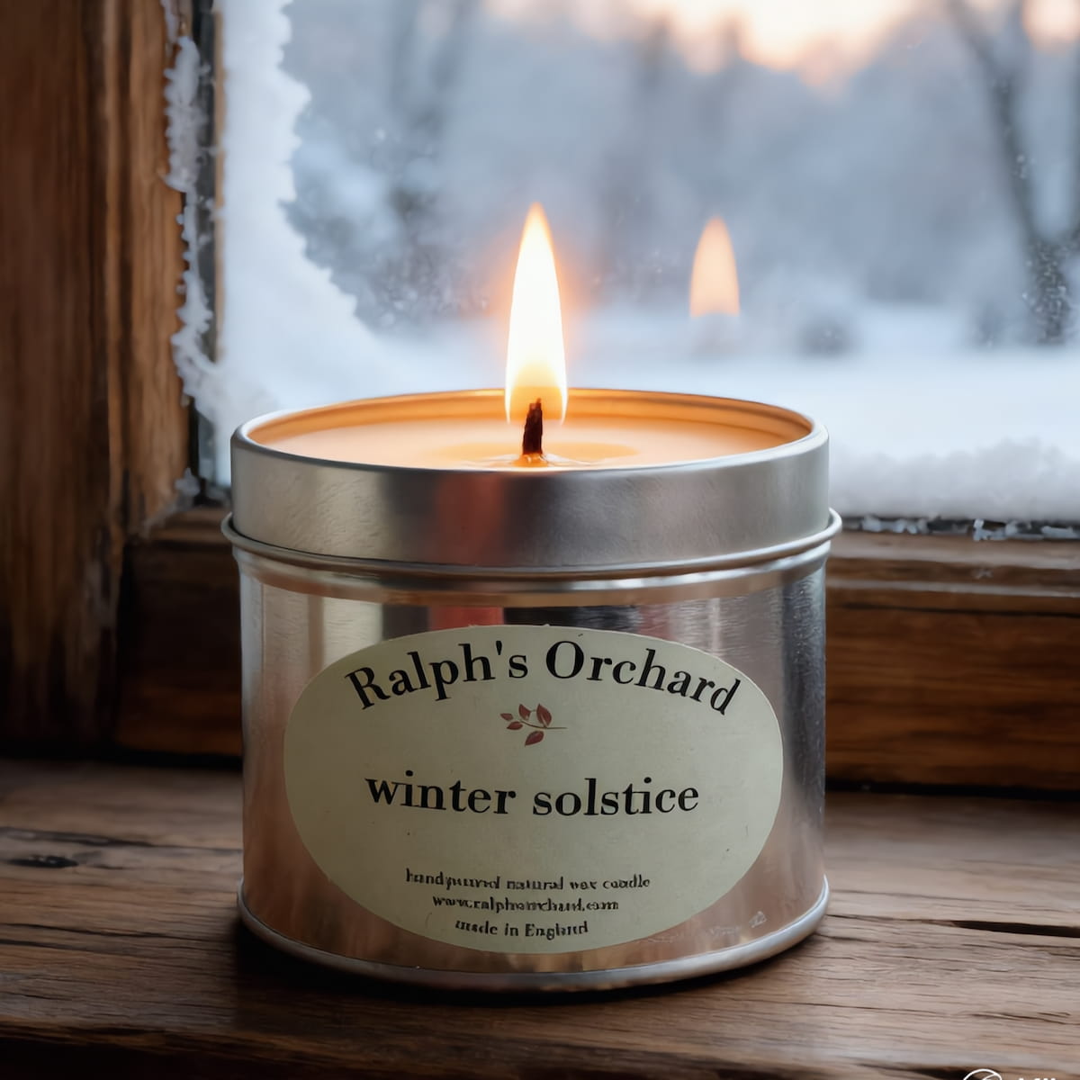Candle in a tin labeled 'Ralph's Orchard winter solstice' on a wooden surface with a snowy window background.