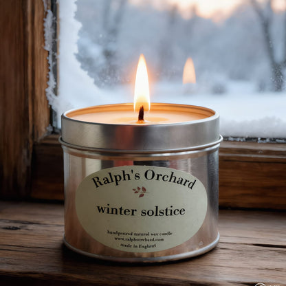 Candle in a tin labeled 'Ralph's Orchard winter solstice' on a wooden surface with a snowy window background.
