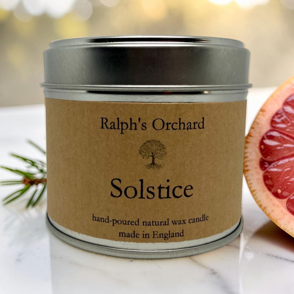 A metal tin candle labeled 'Ralph's Orchard Solstice' sits on a light marble surface with a grapefruit half and pine sprigs beside it. The candle packaging is the main subject, photographed in a styled setting.