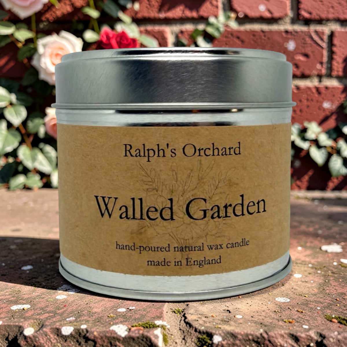 A metal tin candle labeled 'Ralph's Orchard Walled Garden' sits outdoors on a stone surface with a red brick wall and flowers in the background.