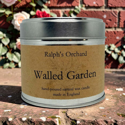 A metal tin candle labeled 'Ralph's Orchard Walled Garden' sits outdoors on a stone surface with a red brick wall and flowers in the background.