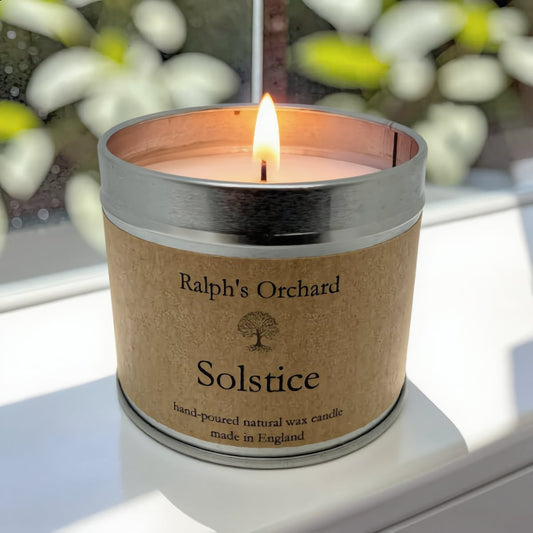 A lit candle in a silver metal tin with a kraft paper label reading 'Ralph's Orchard Solstice hand-poured natural wax candle made in England,' placed on a windowsill with blurred greenery outside.