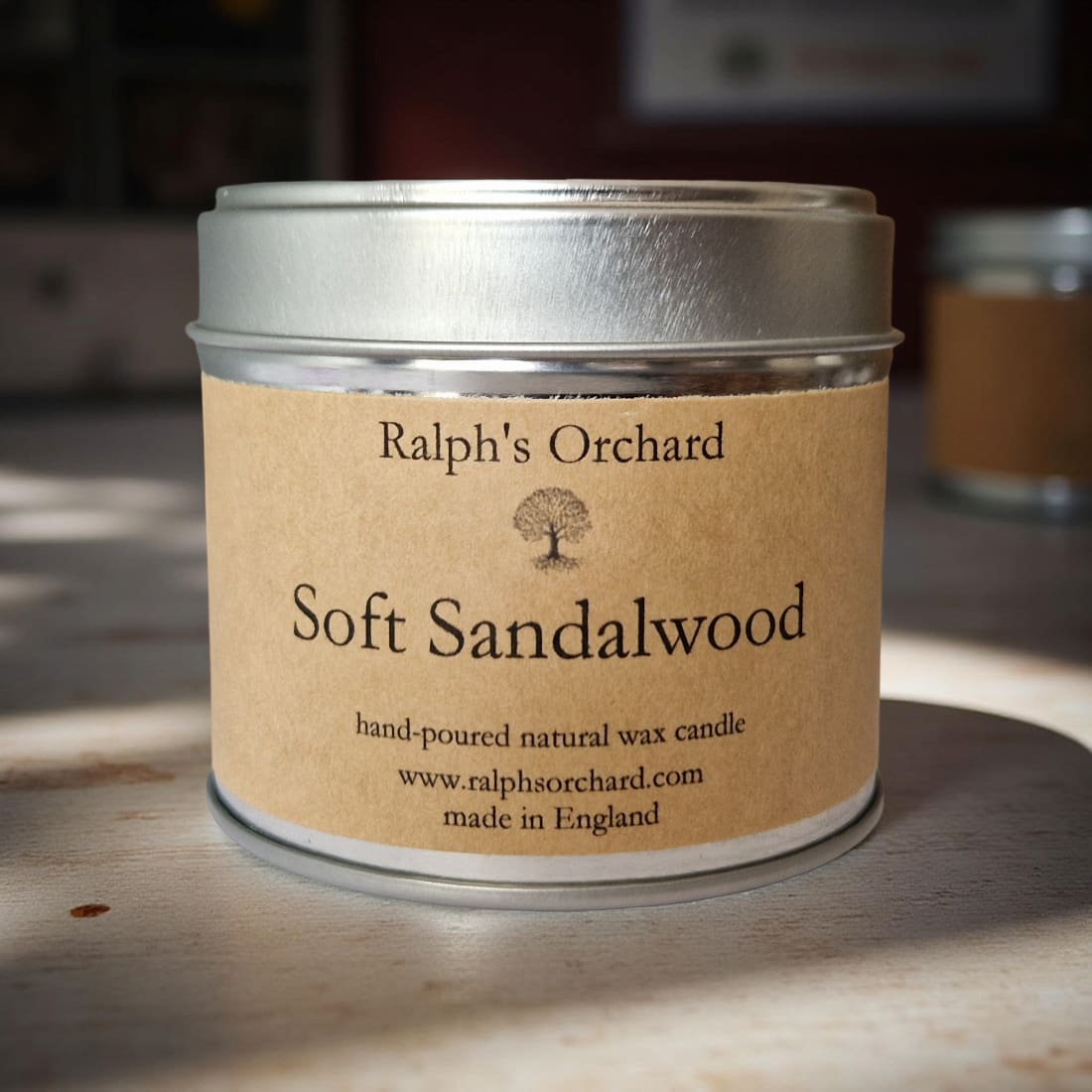 Candle in a tin labeled 'Ralph's Orchard Soft Sandalwood' on a wooden surface.