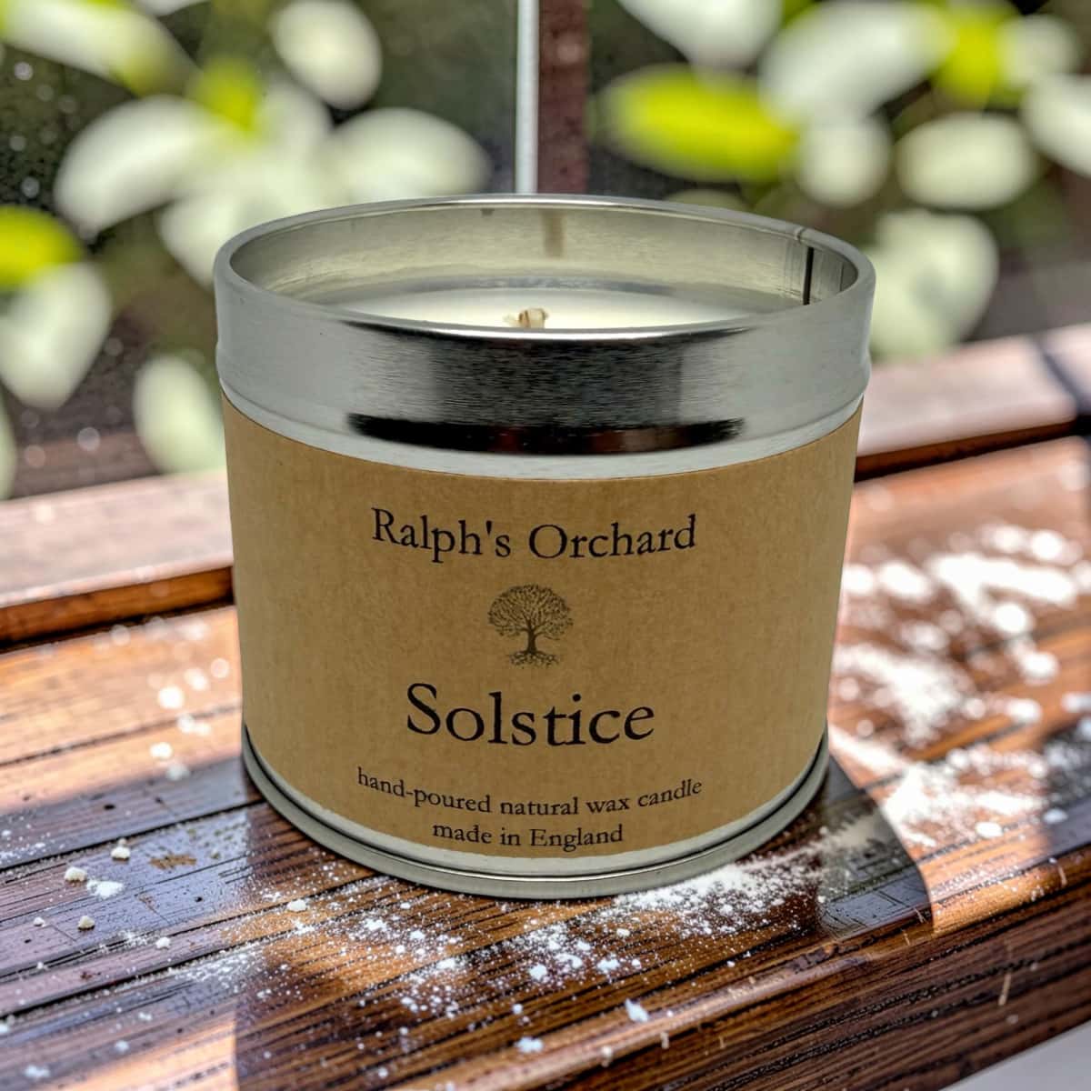 A natural wax candle in a silver metal tin with a kraft paper label reading 'Ralph's Orchard Solstice hand-poured natural wax candle made in England,' placed on a wooden surface outdoors with blurred greenery in the background.