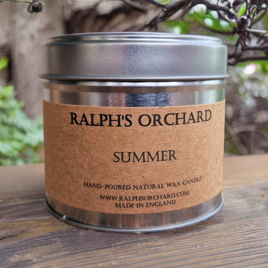 Candle in a metal tin labeled 'Ralph's Orchard Summer' on a wooden surface with a natural background.