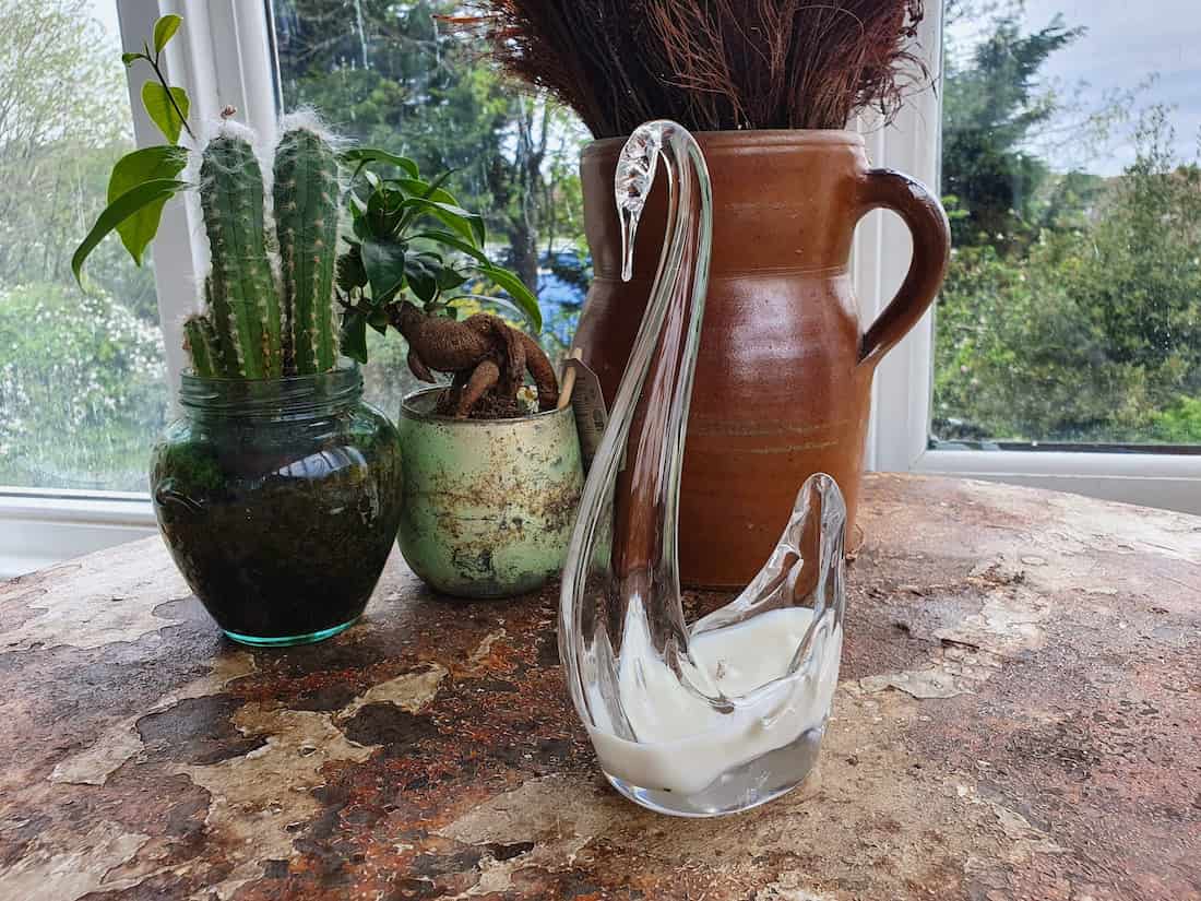 Decorative glass swan on a stone surface with plants and a vase in the background.