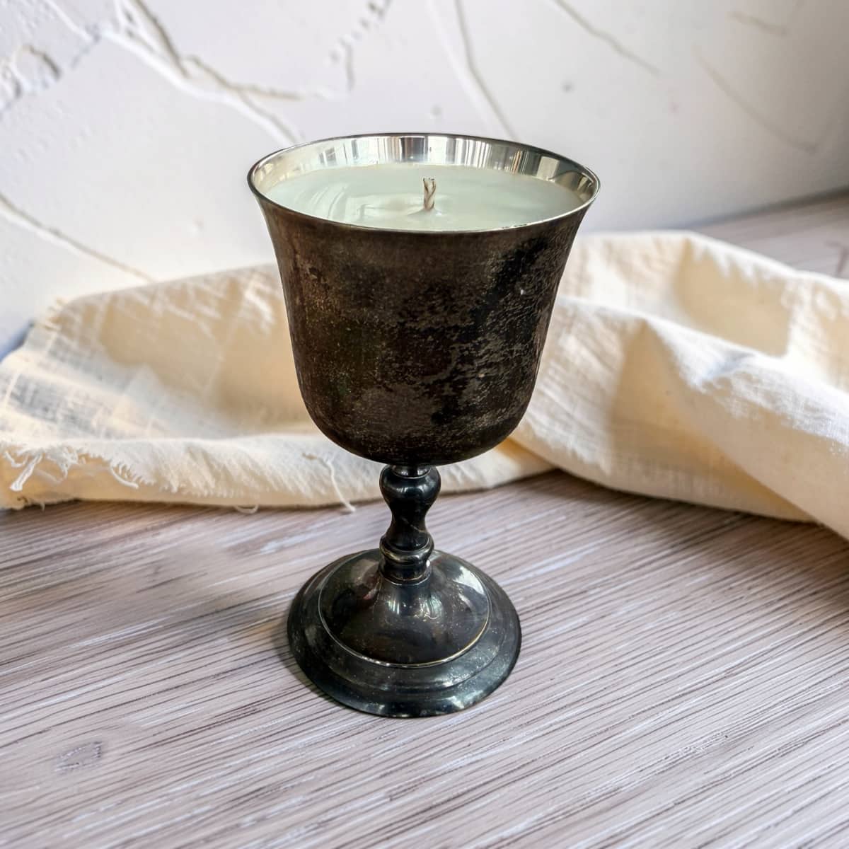 A candle in a dark, textured metal goblet-style holder with a silver rim, set on a light wood surface with a cream fabric napkin and a white textured wall in the background.