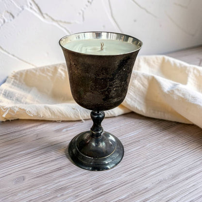 A candle in a dark, textured metal goblet-style holder with a silver rim, set on a light wood surface with a cream fabric napkin and a white textured wall in the background.