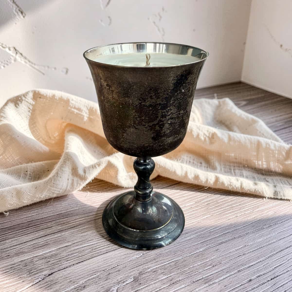 A dark, textured metal candle holder with a single wick, filled with white wax, set on a rustic wooden surface with a draped cream linen cloth in the background.